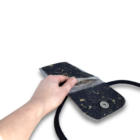 Hand hält die KEDJA Deluxe-Umhängetasche für Mobiltelefone in Schwarz und Grau mit goldenen Akzenten, wobei die Öffnung und die Tasche zu sehen sind.